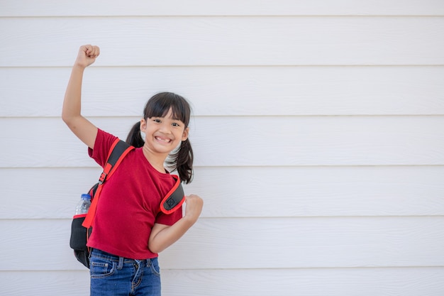 Niña sonriente alegre con mochila grande saltando y divirtiéndose contra la pared blanca. Mirando a la cámara. Concepto de escuela. De vuelta a la escuela