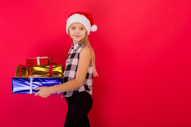 Niña sonriente y alegre con sombrero de Papá Noel con regalo de Navidad en la mano sobre la imagen de fondo rojo