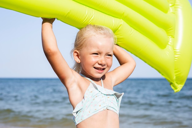 Niña sonriente alegre sosteniendo un colchón inflable en la playa