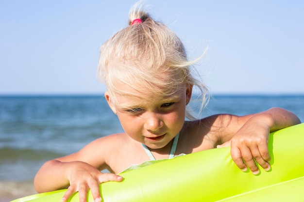 Niña sonriente alegre sosteniendo un colchón inflable en la playa