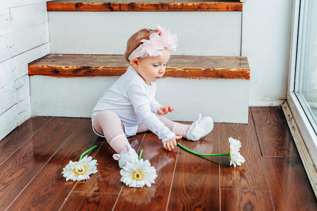 Niña sonriente de un año de edad con corona de primavera sentada en el suelo con luz brillante en la sala de estar cerca de la ventana y jugando con flores de gerbera Niño feliz jugando en casa Concepto de infancia