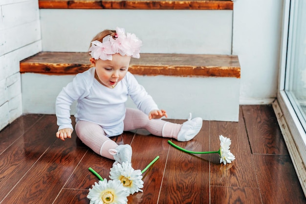 Niña sonriente de un año de edad con corona de primavera sentada en el suelo con luz brillante en la sala de estar cerca de la ventana y jugando con flores de gerbera Niño feliz jugando en casa Concepto de infancia