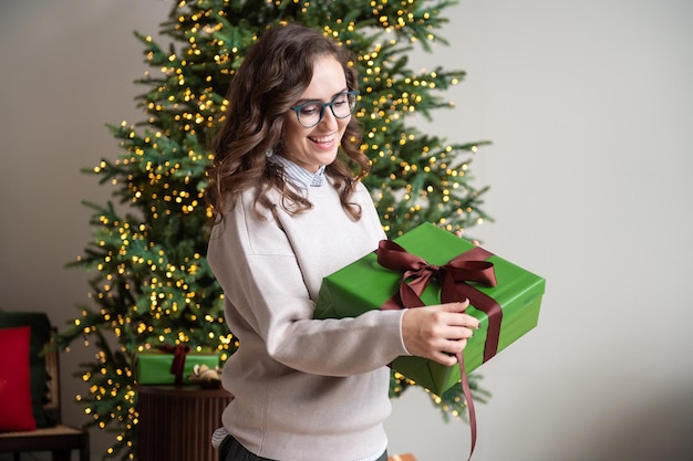 Una niña sonriente con anteojos se regocija en una caja verde con un regalo cerca del árbol de Navidad