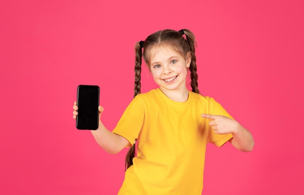 Niña sonriente apuntando al teléfono inteligente en blanco con pantalla negra en la mano