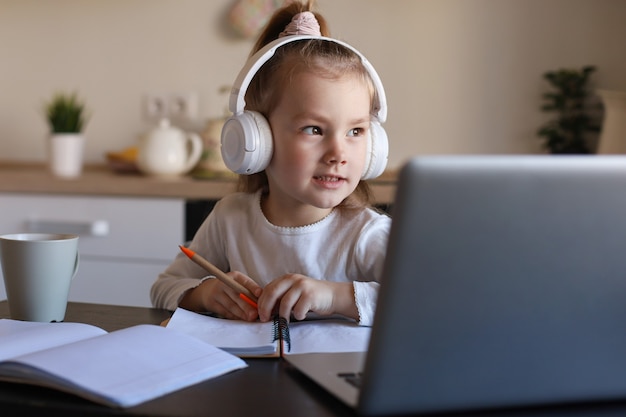 Niña sonriente en auriculares estudio de escritura a mano en línea usando una computadora portátil en casa, un niño pequeño feliz lindo en auriculares toma una lección de Internet o una clase en la PC.