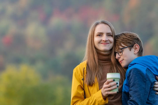 Niña sonriente bebe café o té y abraza a su hijo La madre y el niño cariñosos pasan tiempo juntos caminando por la naturaleza otoñal