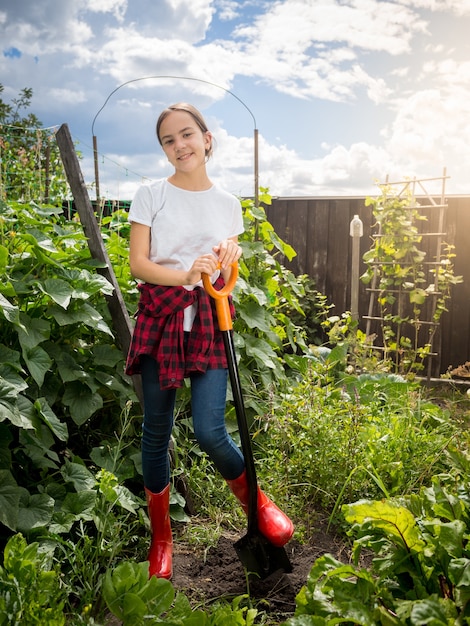 Niña sonriente en botas Wellington sosteniendo una pala y trabajando en el jardín