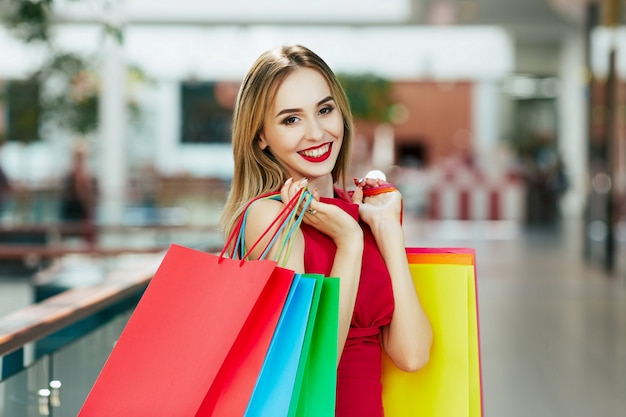 Niña sonriente con cabello castaño claro y labios rojos de pie con coloridos bolsos de compras, concepto de compras, retrato, espacio de copia, emociones felices.