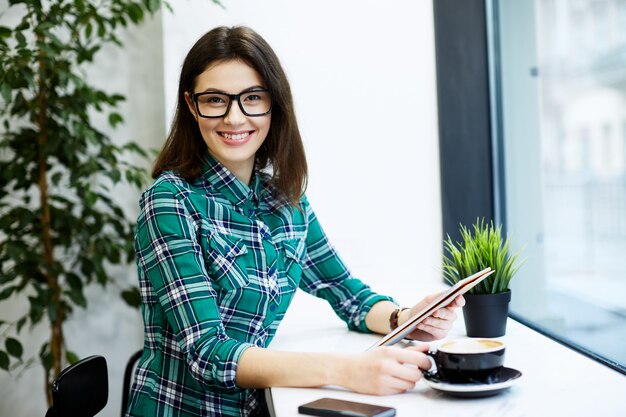 Niña sonriente con cabello negro con camisa y anteojos sentado en la cafetería con tableta y taza de café, concepto independiente, retrato, mirando a cámara.