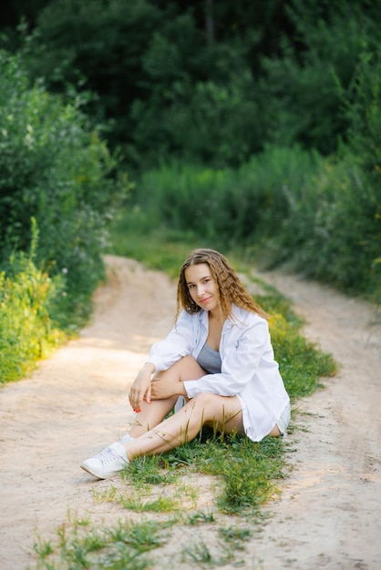 Una niña sonriente con una camisa blanca está sentada en un sendero forestal en un parque verde de verano