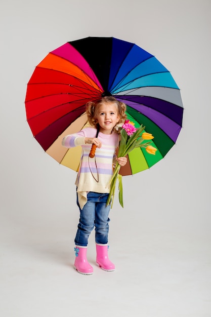 Niña sonriente en camisa rosa a juego y botas de lluvia con paraguas arco iris sobre fondo blanco.