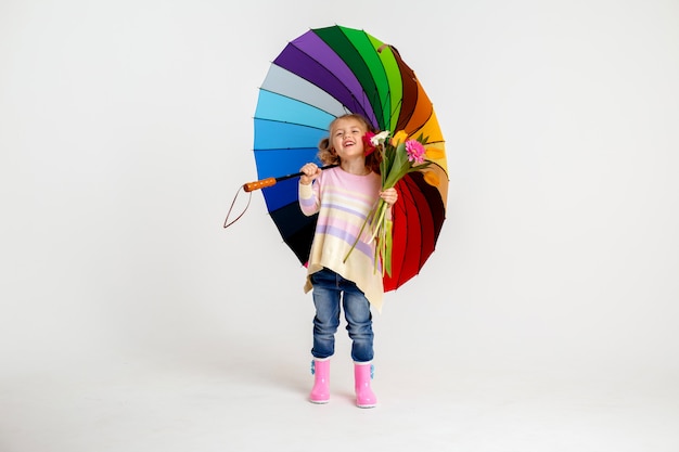 Niña sonriente en camisa rosa a juego y botas de lluvia con paraguas arco iris sobre fondo blanco.