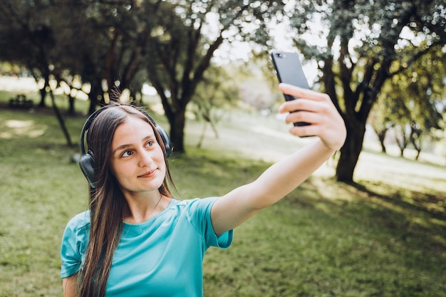 Niña sonriente con una camiseta turquesa y auriculares, tomándose un selfie con un móvil en la naturaleza