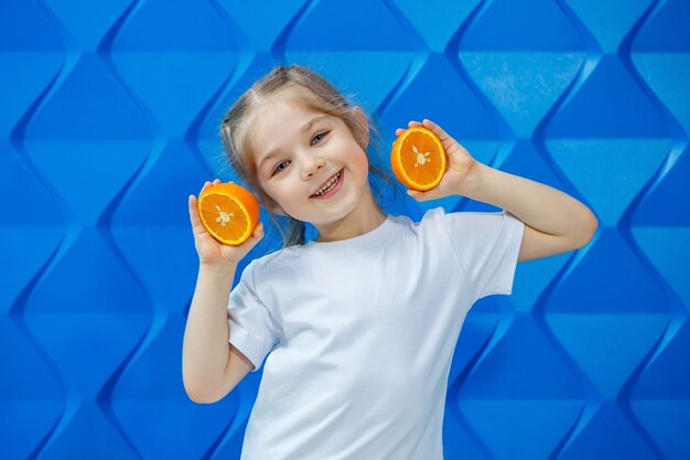 Niña sonriente con coletas en una camiseta blanca sobre un fondo azul con una naranja cortada en sus manos. Emociones infantiles, diversión