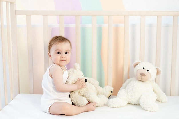 Niña sonriente en una cuna con osos de peluche en una guardería de dormitorio brillante con un traje blanco