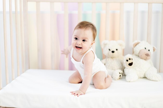 Niña sonriente en una cuna con osos de peluche en una guardería de dormitorio brillante con un traje blanco