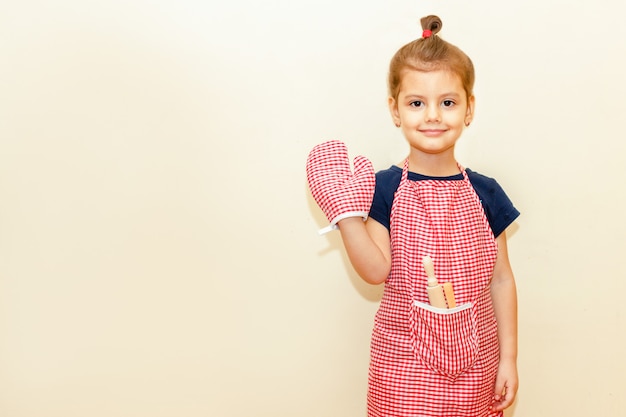 Foto niña sonriente con delantal de chef y agarradera, rodillo de madera y una cuchara sobre fondo beige