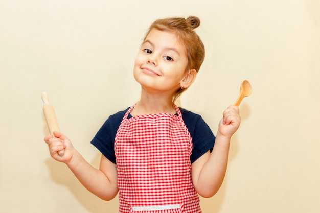 Foto niña sonriente con delantal de chef con rodillo de madera y una cuchara sobre fondo beige