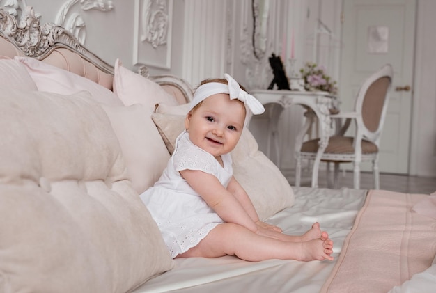 Una niña sonriente con una diadema blanca y un mono está sentada en una cama en una habitación con un hermoso interior