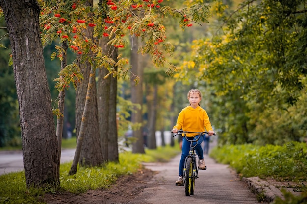 Niña sonriente de diez años con un suéter amarillo monta en bicicleta en la calle de otoño bajo las ramas de un árbol de serbal rojo.