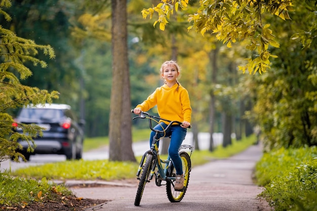 Niña sonriente de diez años en un suéter amarillo monta bicicleta en la calle de otoño bajo las ramas de un árbol de serbal rojo