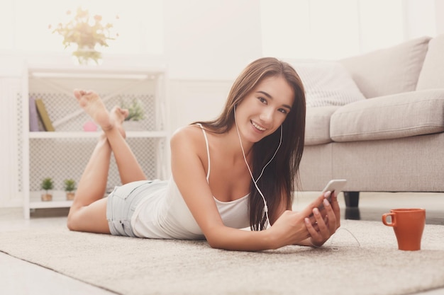 Niña sonriente escuchando música en línea en un teléfono inteligente con auriculares, tendida en el suelo con café en casa, copiando espacio