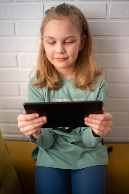 Una niña sonriente está sentada en el reposabrazos del sofá y disfruta jugando en línea en una tableta digital