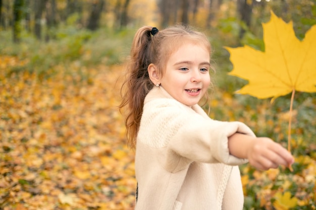 niña sonriente y feliz con abrigo y vestido sosteniendo hojas de otoño, divirtiéndose en el bosque de otoño
