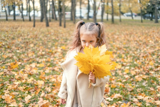 niña sonriente y feliz con abrigo y vestido sosteniendo hojas de otoño, divirtiéndose en el bosque de otoño