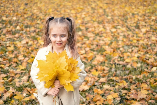 niña sonriente y feliz con abrigo y vestido sosteniendo hojas de otoño, divirtiéndose en el bosque de otoño