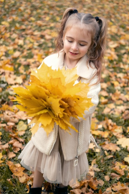 niña sonriente y feliz con abrigo y vestido sosteniendo hojas de otoño, divirtiéndose en el bosque de otoño