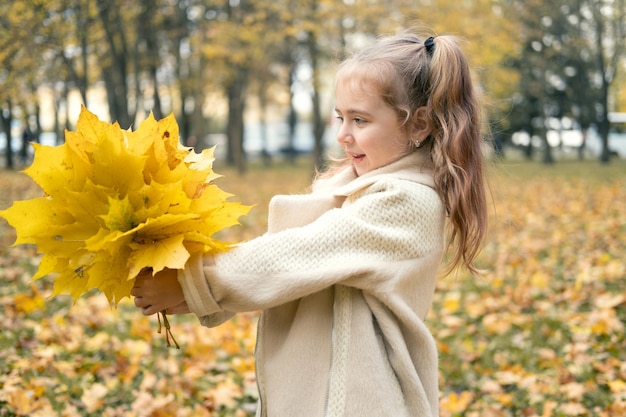 niña sonriente y feliz con abrigo y vestido sosteniendo hojas de otoño, divirtiéndose en el bosque de otoño