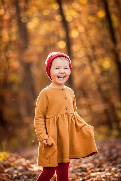 Una niña sonriente feliz en el gorro de punto rojo caminando en el bosque de otoño