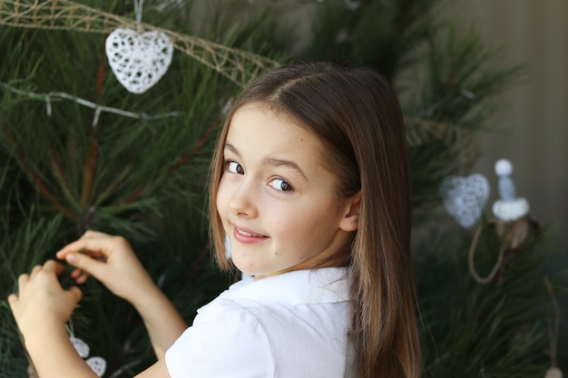 Niña sonriente feliz hermosa que adorna las decoraciones blancas del árbol de navidad que miran