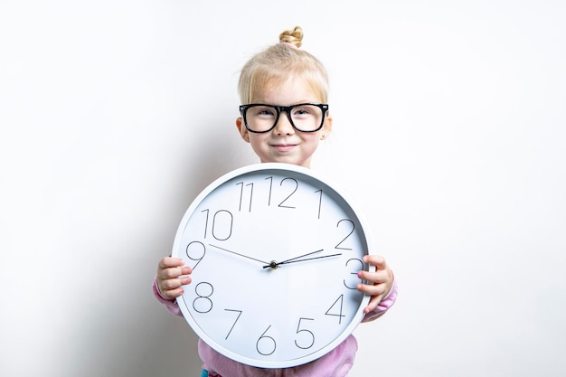 Una niña sonriente con gafas sostiene un gran reloj de pared sobre un fondo claro.