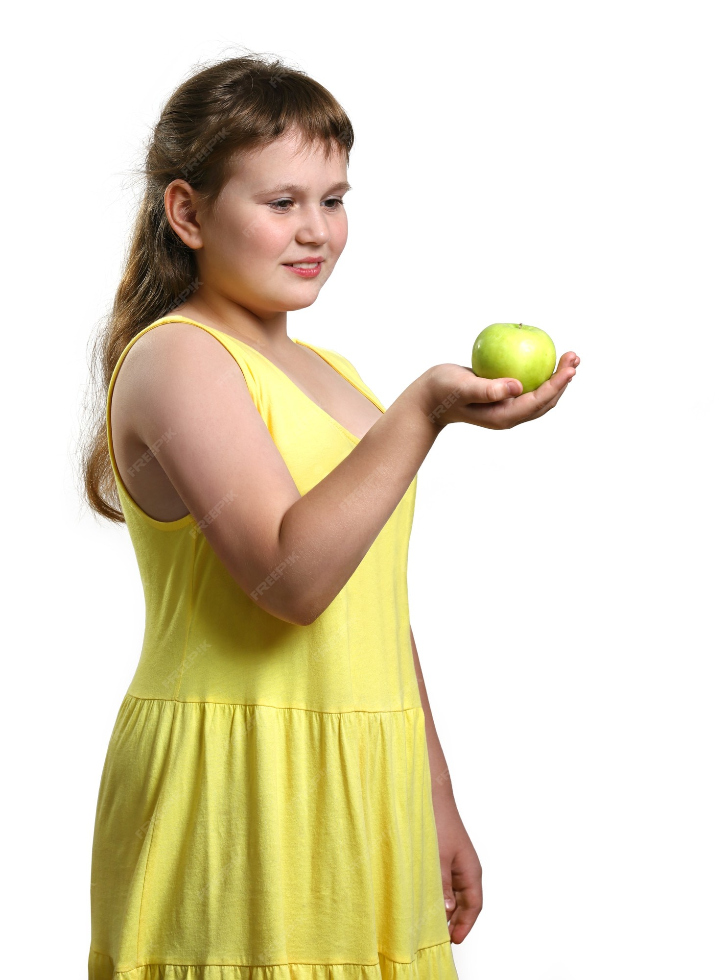 Niña sonriente gordita en vestido amarillo tiene verde en mano y lo mira | Foto Premium