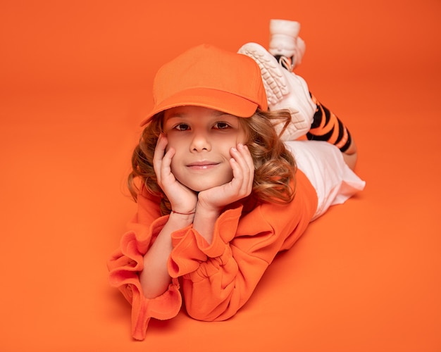 Niña sonriente con una gorra y ropa de moda acostada en el piso en estudio sobre fondo naranja. Bosquejo