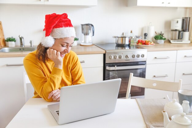 Niña sonriente con gorro de Papá Noel con videollamadas familiares por webcam. Mujer con laptop sentada en la cocina en casa usando chat de reunión virtual en vacaciones. Feliz navidad y año nuevo nuevo normal.