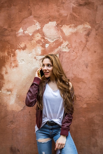 Foto niña sonriente hablando con su teléfono inteligente con una pared roja en el fondo en vertical