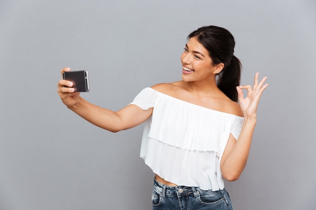 Niña sonriente haciendo foto selfie en smartphone y mostrando gesto bien aislado en una pared gris