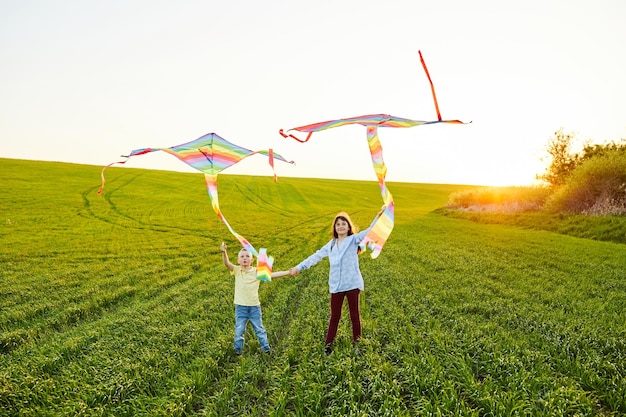 Niña sonriente y hermano niño de pie y tomados de la mano con cometas de colores voladoras en el prado de hierba alta Momentos felices de la infancia