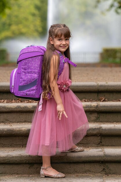 Una niña sonriente con un hermoso vestido rosa y pelo largo con una mochila en los escalones del par