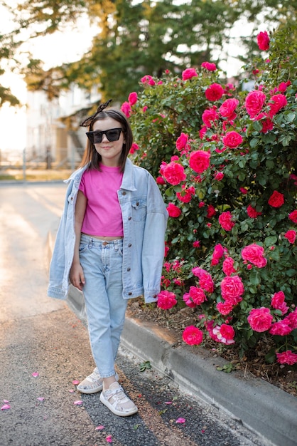 Una niña sonriente y linda usa una chaqueta de mezclilla de mamá y gafas de sol posando sobre flores rosas rosadas