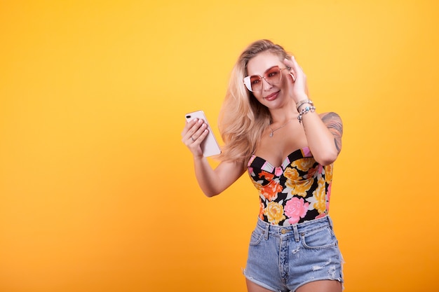 Niña sonriente mirando la pantalla del teléfono en sus manos con gafas de sol en estudio sobre fondo amarillo