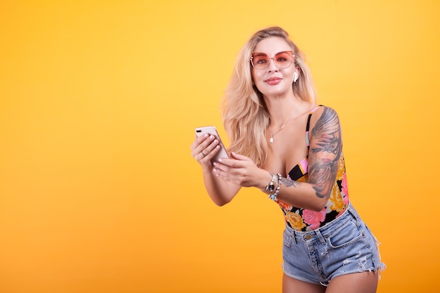 Niña sonriente mirando la pantalla del teléfono en sus manos con gafas de sol en estudio sobre fondo amarillo