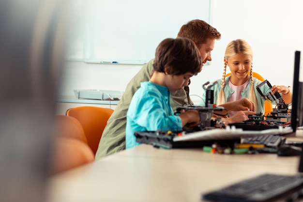 Niña sonriente y niño concentrado construyendo robots usando juegos de construcción sentados con su maestro en su lección de ciencias