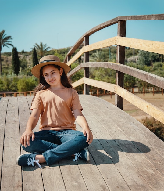 Una niña sonriente niño sentado con las piernas cruzadas en el puente de madera en el parque en un día soleado con sombrero de paja, camiseta naranja y jeans y mirando directamente a la cámara