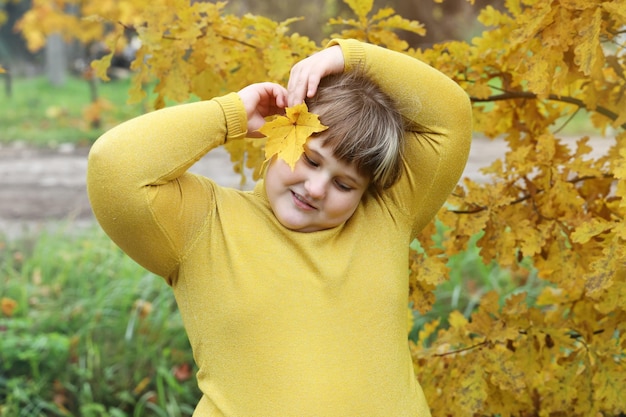 Niña sonriente obesa se para al aire libre y coloca una hoja de arce amarilla para la oreja derecha