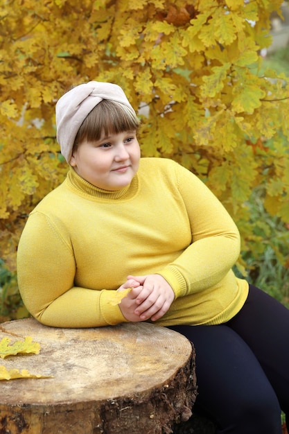 Niña sonriente obesa se sienta en tocón de madera cerca de árboles otoñales con follaje amarillo