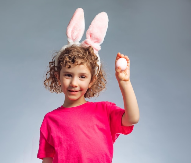 La niña sonriente con orejas de conejo sosteniendo un huevo de Pascua La niña con el pelo rizado se encuentra sobre un fondo gris